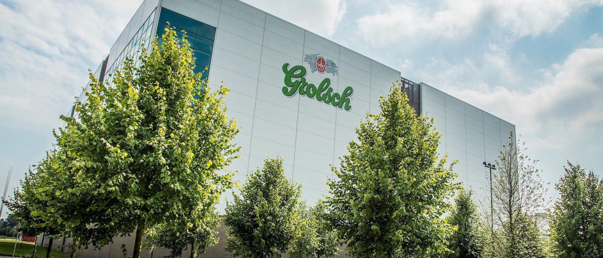 Buitenkant van de Grolsch-brouwerij met het groene logo op de gevel, achter bomen.