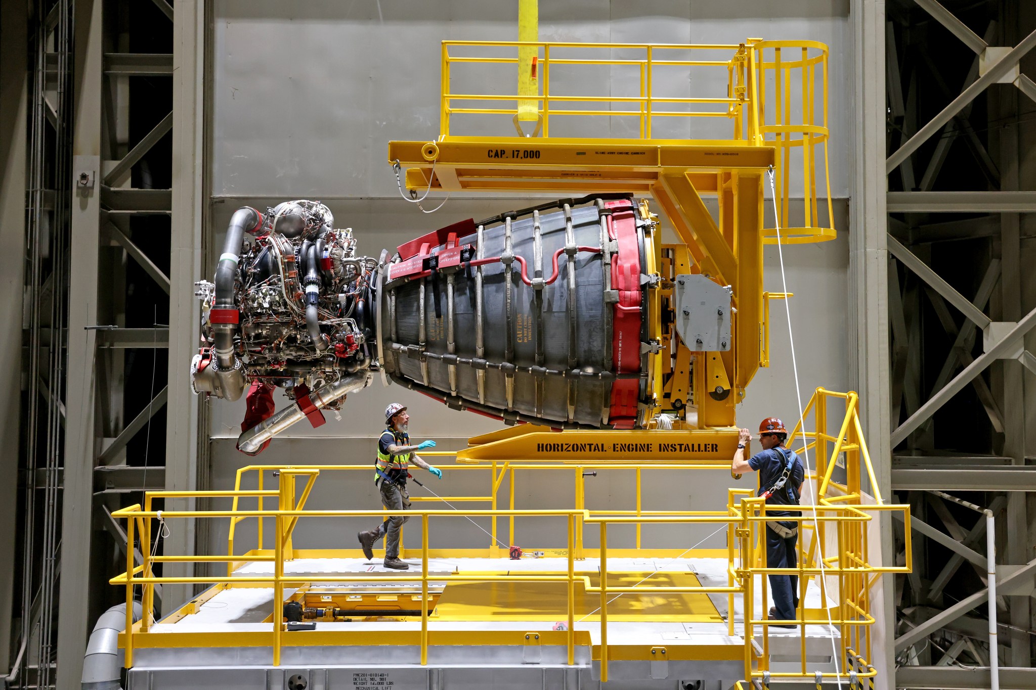 NASA-foto uit 2023: twee technici staan bij een horizontaal opgehangen RS-25-raketmotor in een gele installatiehal.