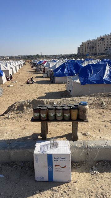 Een WFP-voedselpakket en potten met eten op de voorgrond, met daarachter een kamp met tenten.